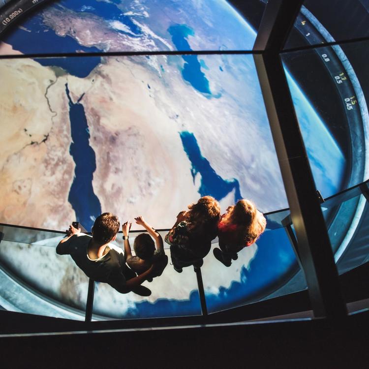 Een familie met twee kinderen staat op een glazen reling en kijkt uit over een grote interactieve wereldbol in het Discovery Museum